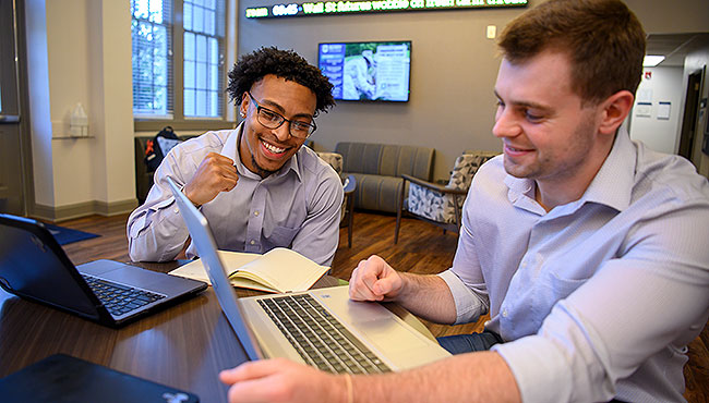 Male students working on laptops in the Campbell School of business with Wall Street ticker in the background