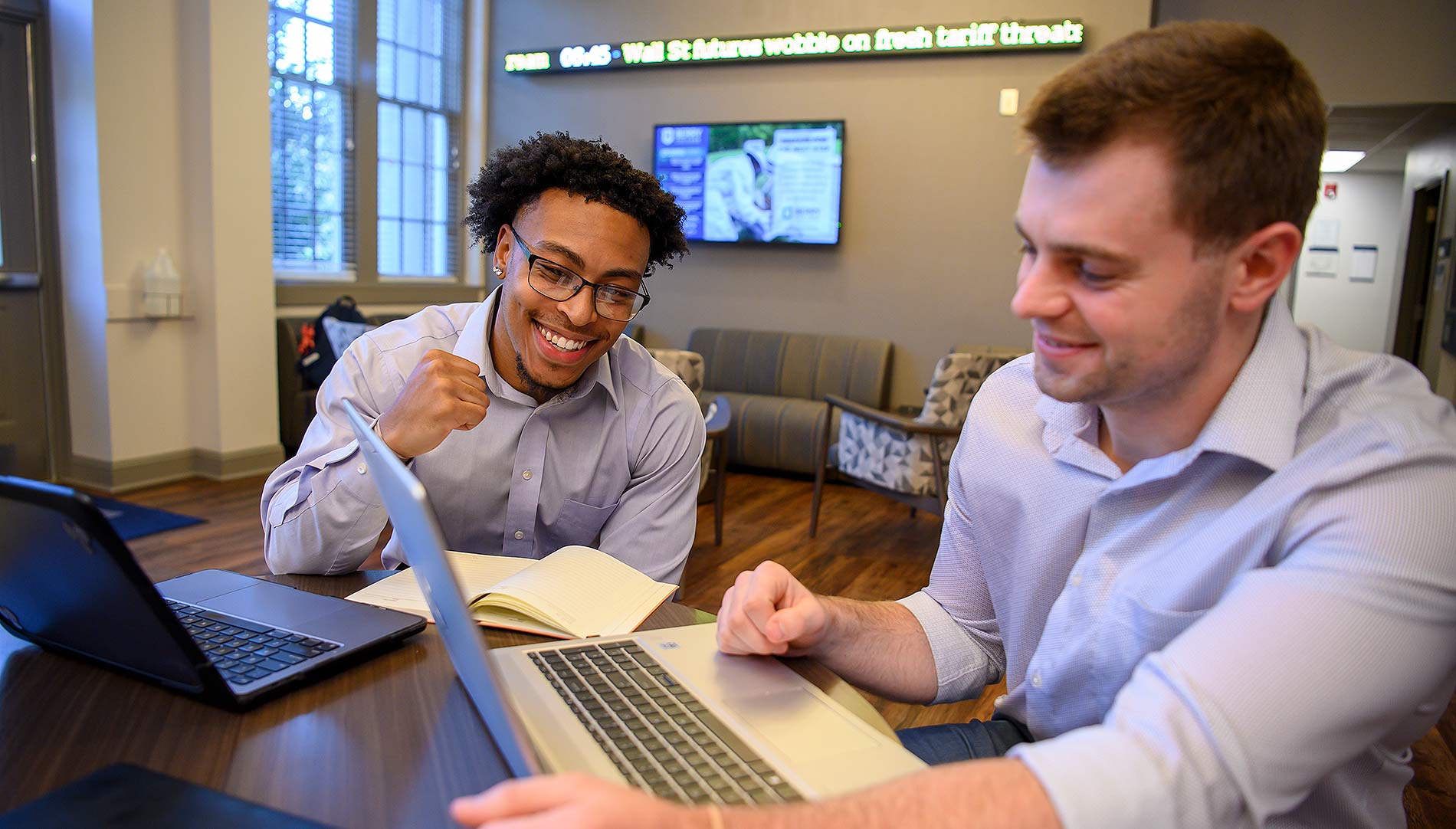 Male students working on laptops in the Campbell School of business with Wall Street ticker in the background
