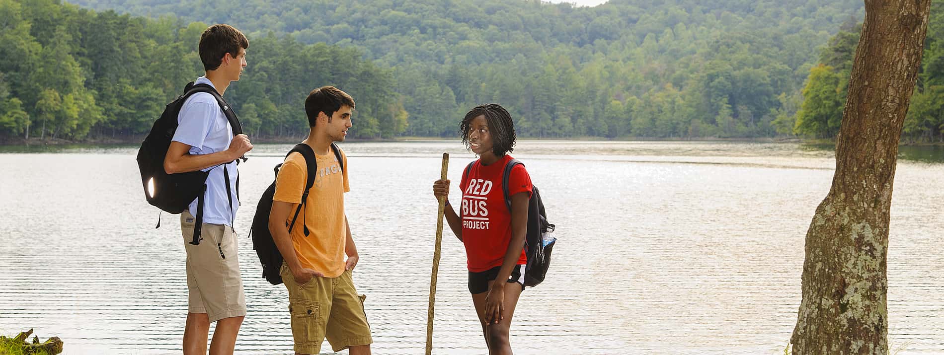 Three students with backpacks and walking sticks standing by a lake