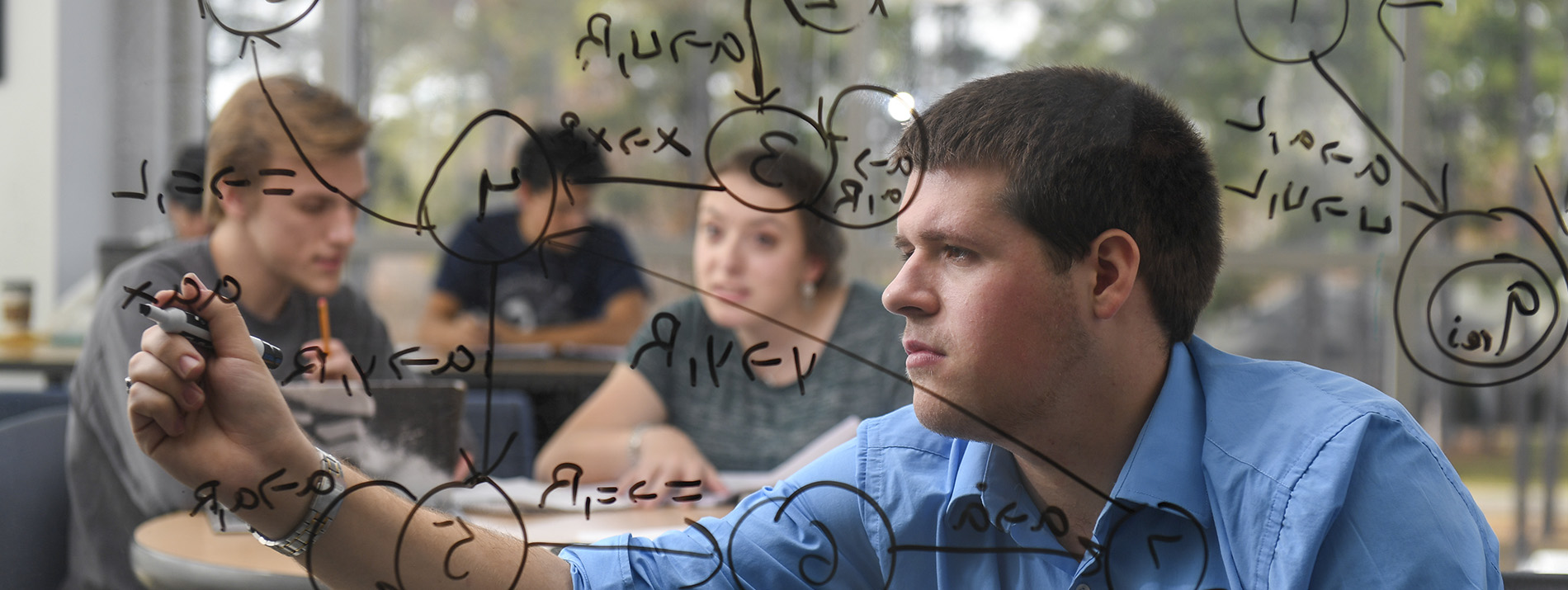 student writing on glass board with marker while other students look on from the background